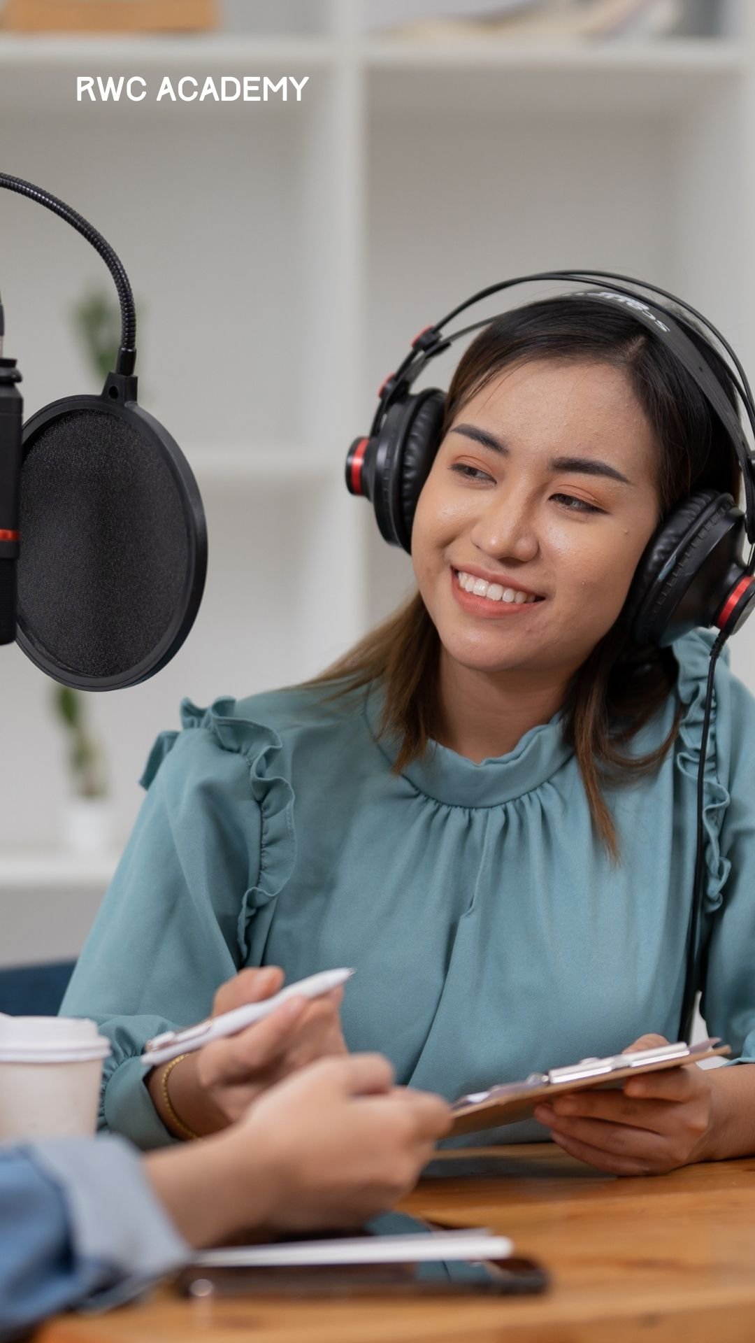 A smiling woman wearing headphones speaks into a professional microphone while holding a clipboard, suggesting she is recording a podcast or participating in an interview.